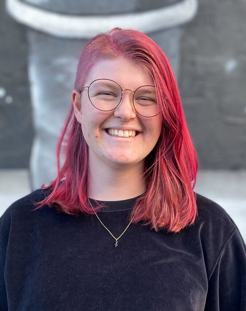 Alexa, who has shoulder-length pink hair and wire-rimmed glasses, stands in front of a grey brick wall